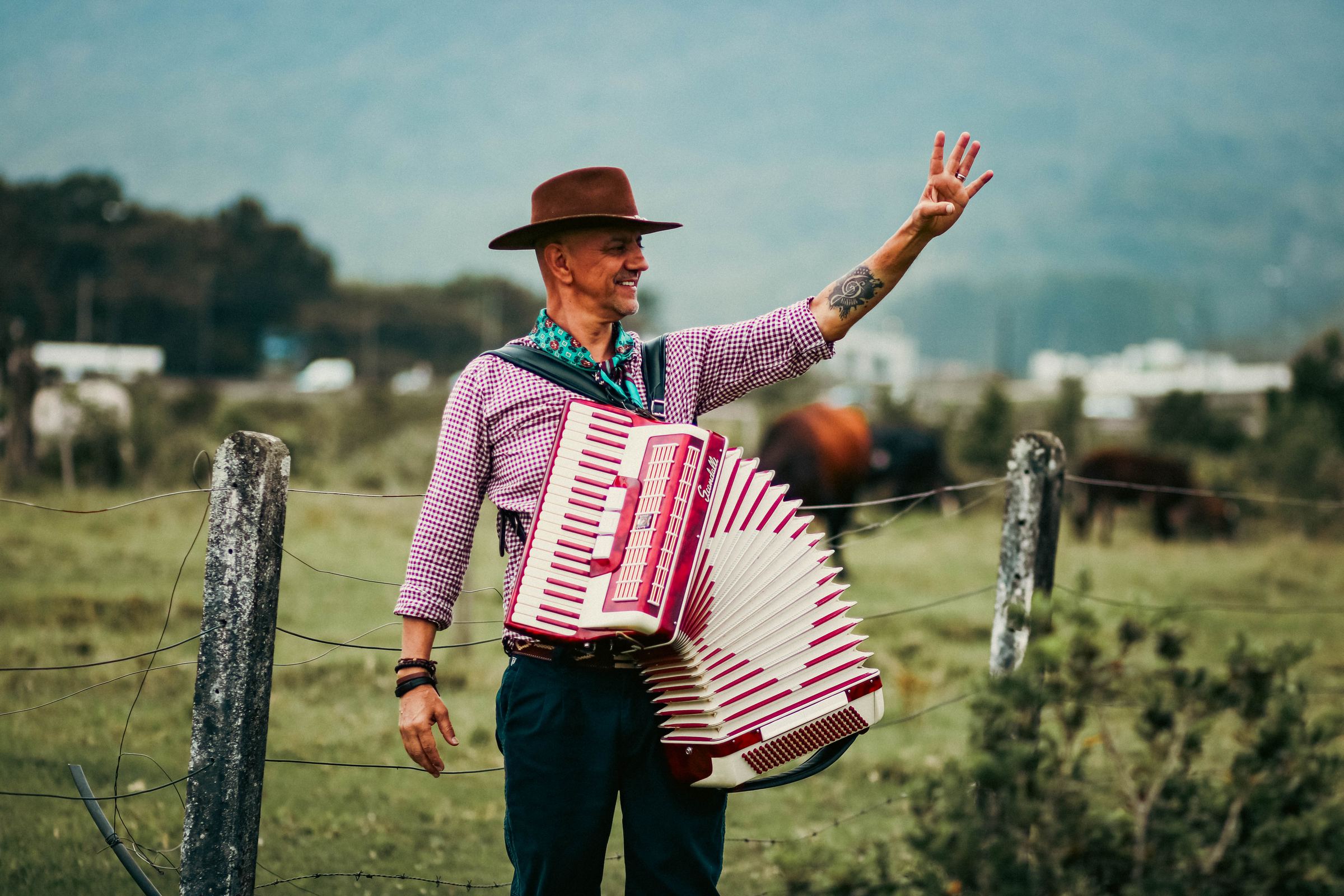 Carlos Magrão com a gaita vermelha no campo, acenando, com as montanhas ao fundo