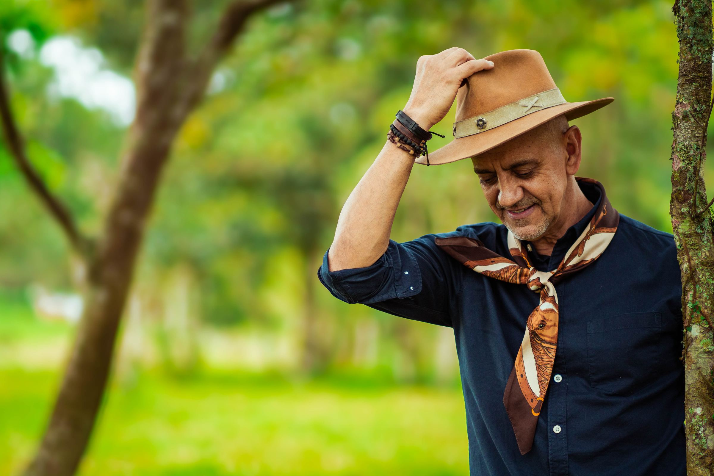 Carlos Magrão sorrindo, com a mão no chapéu, entre as árvores