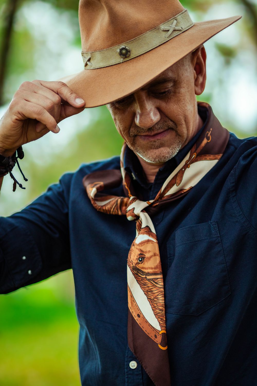 Carlos Magrão sorrindo, de chapéu e lenço, entre as árvores